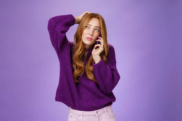 頭を引っかく携帯電話を介して予約をするかわいい赤毛の女性を混乱させ、思考の選択や記憶として検索し、紫色の壁の上にスマートフォンと話す — ストック写真