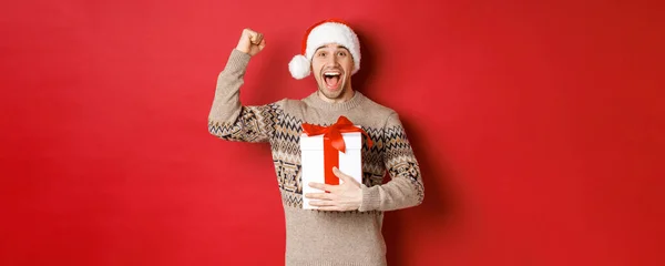 Imagem de homem bonito feliz e animado, reicing presente de Natal, levantando as mãos em triunfo e sorrindo, celebrando o ano novo, de pé sobre fundo vermelho em Papai Noel — Fotografia de Stock