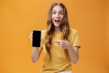 Harika akıllı telefon özellikleriyle şaşırmış bir kız. Cep telefonunun uygulama ekranını işaret ettiğini gösteren dalgalı doğal saç stiline sahip neşeli ve şaşkın Avrupalı ince kadının portresi