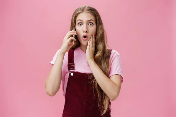 Vita-up shot di donna scioccata imparare notizie mozzafiato tramite smartphone parlando sul telefono cellulare in possesso di orecchie labbra pieghevoli e ansimando a sorpresa spuntando gli occhi davanti alla fotocamera preoccupati per il muro rosa — Foto Stock