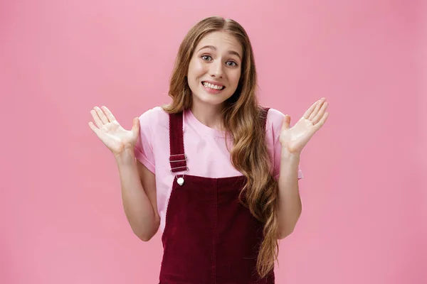 Lo siento, no tengo idea. Tonto ignorante linda joven estudiante de aspecto amistoso en dungarees sobre camiseta encogiéndose de hombros con las palmas levantadas en rendición sonriendo incómodo como si no tuviera idea sobre la pared rosa — Foto de Stock