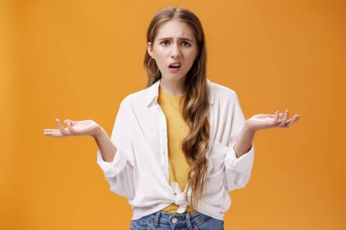 What, I do not understand. Portrait of arrogant questioned impolite girl being pissed with questiones shrugging with hands spread sideways making irritated grimace standing confused over orange wall