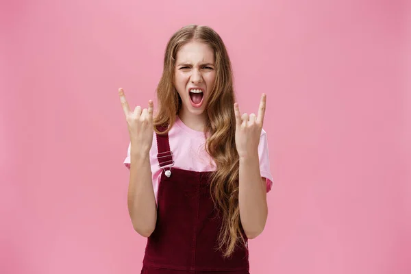 Cute and excited party girl with wavy hairstyle in corduroy overalls yelling yeah and showing rock-n-roll gesture having fun enjoying hard music being daring and rebellious over pink wall — Stock Photo, Image
