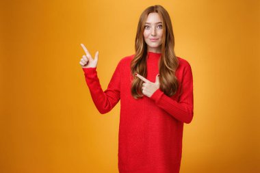 Yumuşak ve nazik, kızıl saçlı, kırmızı, sıcak kazaklı, mutlu bir şekilde gülümseyerek sol üst köşeyi gösteren, ilginç bir yer gösteren, turuncu arka plan üzerinden ziyaret etmeyi öneren kız.