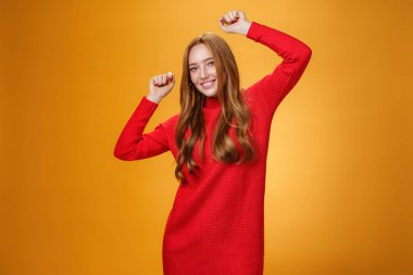 Neşeli ve kaygısız, rahat, kırmızı, sıcak elbiseli, parti veren, havalı müziğin tadını çıkaran, elleri havada, turuncu arka planda kameraya gülümseyen bir kız.