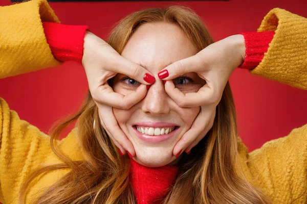 Menina ruiva gosta de brincar e fingir que ela criança novamente fazendo máscara engraçada com os dedos no rosto como super-herói sorrindo amplamente, tolo em torno brincalhão sobre fundo vermelho — Fotografia de Stock