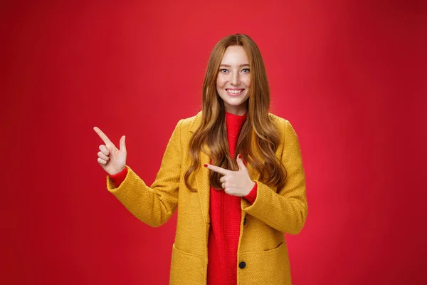 Χαρούμενα φιλικό αναζητούν και ενεργοποιημένο χρήσιμο κορίτσι κοκκινομάλλα σε κίτρινο παλτό δείχνει τον τρόπο που δείχνει στην πάνω αριστερή γωνία και χαμογελώντας γενικά με ικανοποιημένη χαμόγελο ποζάροντας ευχαριστημένος πάνω από το κόκκινο φόντο — Φωτογραφία Αρχείου