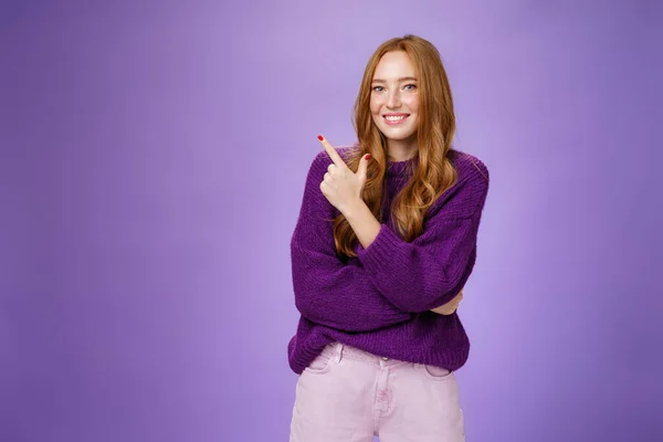 Encantadora mujer pelirroja brillante de aspecto amigable con pecas y maquillaje en suéter cálido púrpura apuntando a la esquina superior izquierda y sonriendo encantado y lindo en la cámara como muestra lugar fresco pasar el rato —  Fotos de Stock