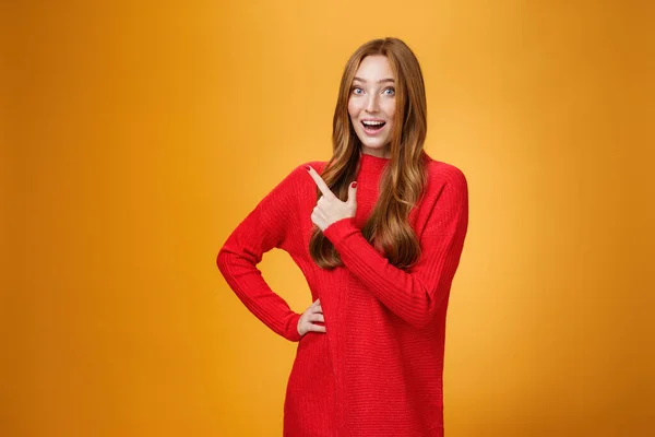 Energizado otimista e alegre menina de gengibre caucasiano de aparência amigável em camisola vermelha apontando para o canto superior esquerdo com sorriso animado e alegre entusiasmado posando encantado contra fundo laranja — Fotografia de Stock