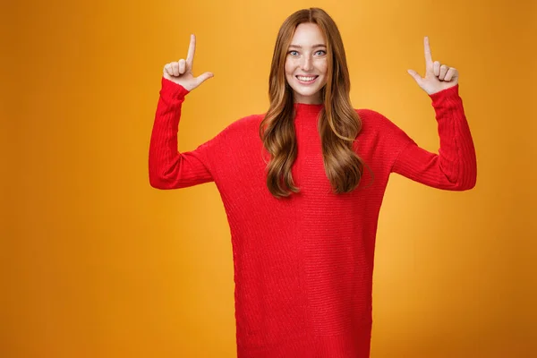 Entusiasta y excitada pelirroja enérgica en suéter rojo cálido apuntando hacia arriba con las manos levantadas y sonriendo ampliamente emocionada con la promoción impresionante, sintiéndose asombrada contra el fondo naranja —  Fotos de Stock