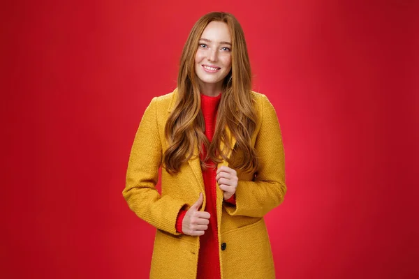 Stylisch und niedlich gut aussehende rothaarige Frau in gelbem Mantel auf dem Weg zur Arbeit, ergreift Kaffeebestellung mit niedlichem freundlichem Lächeln Knopf anrührend und posiert über rotem Hintergrund — Stockfoto
