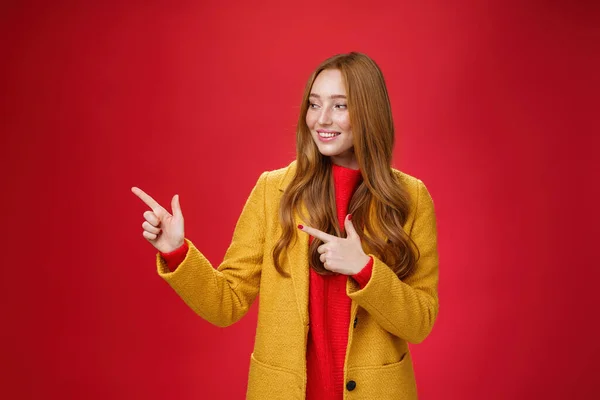 Ritratto di donna rossa attraente curiosa e felice soddisfatta in cappotto giallo che punta e guarda a sinistra con un sorriso incuriosito che osserva uno spazio di copia impressionante sullo sfondo rosso — Foto Stock
