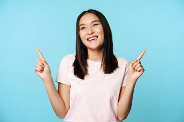 Gülümseyen esmer Asyalı kadın, iki tarafı da gösteriyor ve gülüyor, mağazayı seçiyor, mavi arka planda duruyor.