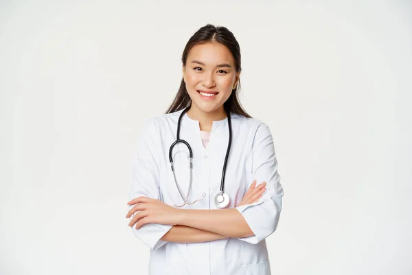 Zdravotní a lékařská koncepce. Usmívající se asijský lékař v uniformě, hledá jistotu na kameru, léčení pacientů, stojící nad bílým pozadím — Stock fotografie
