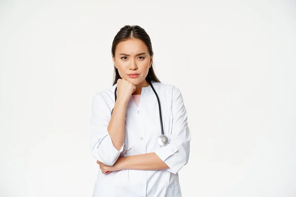 Image of angry nurse, asian female doctor looking annoyed and bothered, furrow eyebrows and sulking thoughtful, standing over white background — Stock Photo, Image