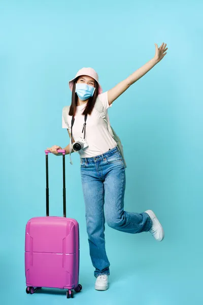 Felice ragazza asiatica in maschera medica, turista in posa eccitata con valigia, andare in vacanza, viaggiare all'estero, in piedi su sfondo blu — Foto Stock