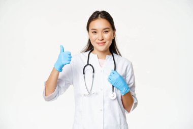 Gülümseyen Asyalı kadın doktor onaylıyor, lastik eldiven giyiyor ve klinik üniforması giyiyor, beyaz arka planda duruyor.
