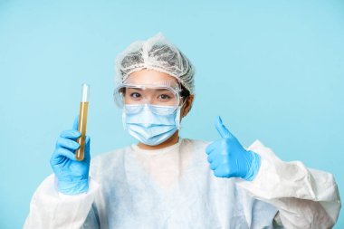 Asyalı kadın doktor, kişisel koruyucu ekipman laboratuvar çalışanı, başparmaklarını gösteriyor ve test tüpü analizi yapıyor, mavi arka planda duruyor.