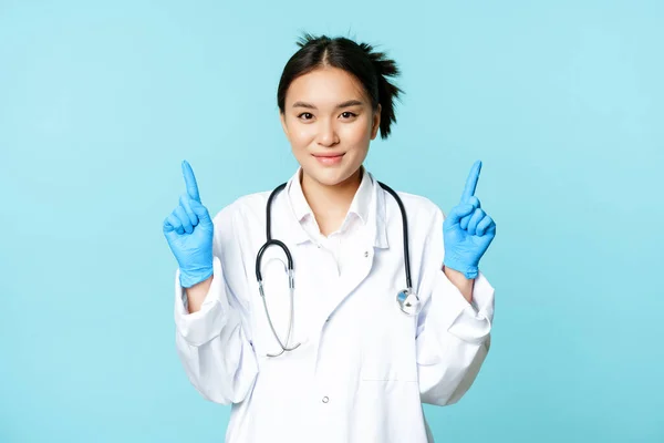 Usmívající se asijský lékař, zdravotní sestra v uniformě, rukavice, ukazující prsty nahoru, ukazující informace o zdravotní péči, stojící nad modrým pozadím — Stock fotografie