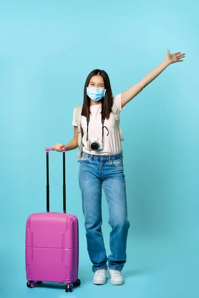 Plná délka šťastné korejky v masce, stojící s kufrem, zvedající ruku zasněný, užívající si dovolenou v zahraničí, stojící nad modrým pozadím — Stock fotografie