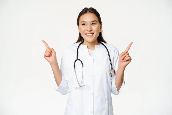 Aziatische vrouw arts in medische uniform en stethoscoop, wijzen zijwaarts, tonen twee producten, beide richtingen, glimlachen blij en kijken naar links, witte achtergrond — Stockfoto