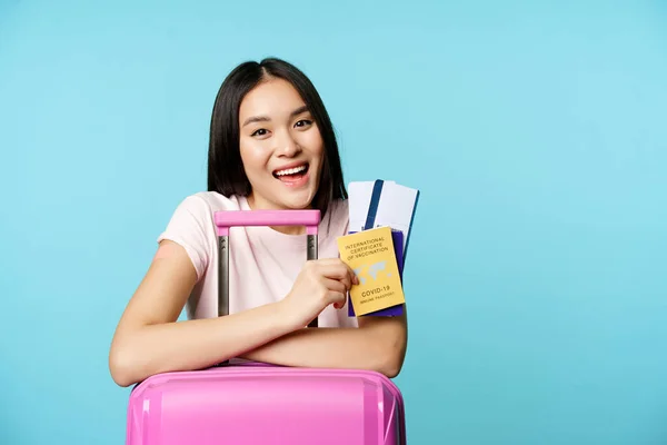 Begeistertes asiatisches Mädchen zeigt Reisepass und Impfpass, lächelt zufrieden, steht mit Koffer, blauer Hintergrund — Stockfoto