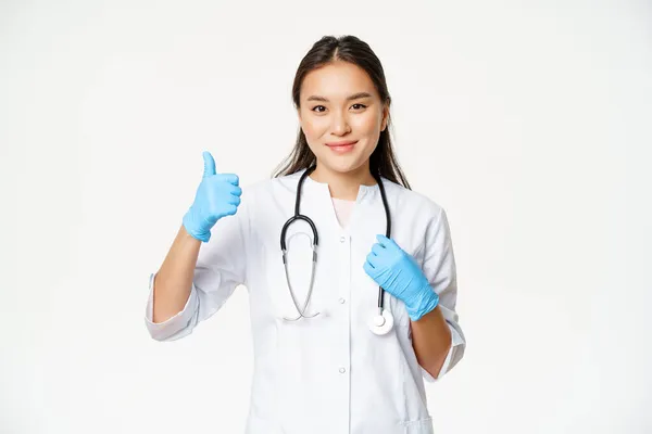 Sonriendo asiático médico muestra los pulgares hacia arriba, lleva guantes de goma y uniforme de la clínica, se para sobre fondo blanco — Foto de Stock