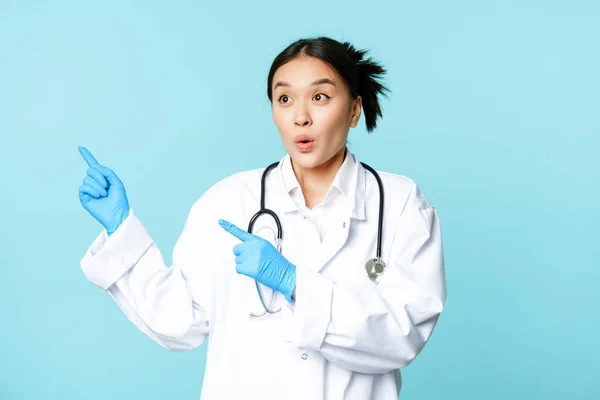 Sorprendido médico mujer asiática o enfermera, señalando los dedos a la izquierda y mirando asombrado en la publicidad, espacio de copia, de pie en uniforme sobre fondo azul — Foto de Stock