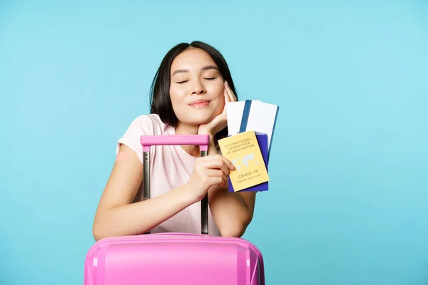 Sorridente turista asiatica sognante, ragazza pensa di viaggiare, in possesso di passaporto e biglietti, coronavirus certificato di vaccinazione internazionale, andare in viaggio all'estero, sfondo blu — Foto Stock