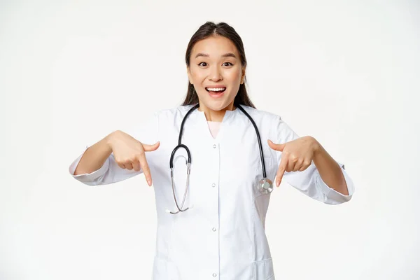 Nadšení zdravotnický personál, asijské ženy zdravotnický pracovník, ukazuje prsty dolů a usmívá ohromeně, ukazuje slevy, prodej na klinice, bílé zázemí — Stock fotografie