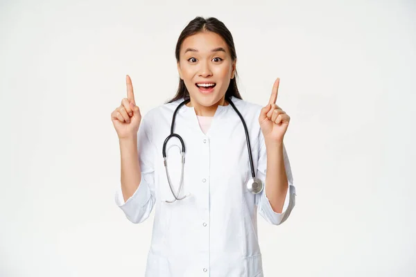 Enfermera sonriente emocionada, doctora en uniforme médico señalando hacia arriba, mostrando venta promocional, descuento en la clínica de salud, de pie sobre fondo blanco — Foto de Stock