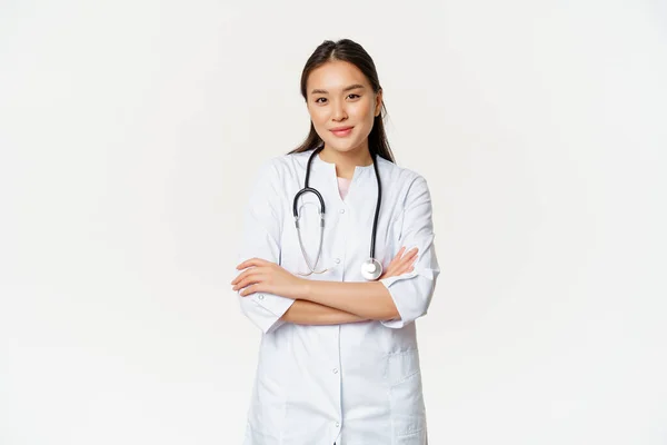 Bild av självsäker kvinnlig läkare, praktikant med stetoskop och medicinsk mantel, kors armar som professionell, ser säker på kameran, vit bakgrund — Stockfoto