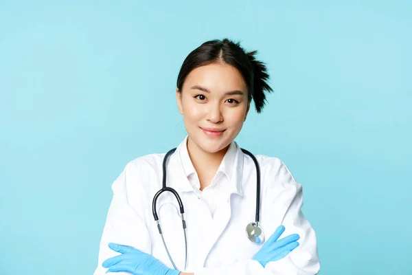 Usmívající se asijky lékař, zdravotní sestra v rukavicích a klinice uniforma, křížové paže a vypadá odhodlaně, modré pozadí — Stock fotografie