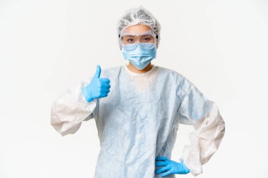 Kişisel koruyucu ekipman ve lastik eldiven takan Asyalı kadın doktor veya hemşire, sağlık maskesi, baş parmak gösterme, aşı ve covid-19 kampanya konsepti
