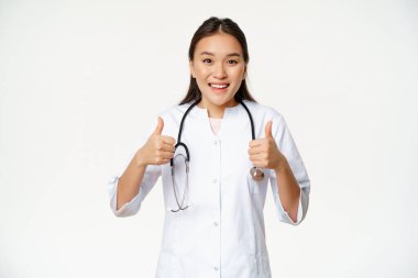 Gülümseyen Asyalı kadın doktor, tıbbi bornoz giyiyor, başparmaklarını kaldırıyor, sağlık ürünleri öneriyor, beyaz arka planda duruyor.