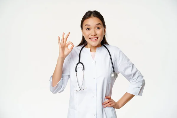 A mosolygó ázsiai orvos szerint rendben van a jel, orvosi köpenyt visel. Női kórházi dolgozó egyenruhában ajánlja smth, álló felett fehér háttér — Stock Fotó