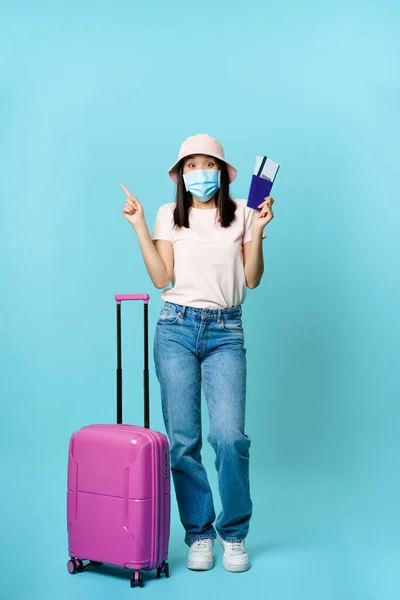 Vertikální záběr šťastné korejky v lékařské masce, s letenkami a pasem, stojící s kufrem, snění o budoucí dovolené, modré pozadí — Stock fotografie