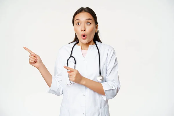 Πορτρέτο της ενθουσιώδης γυναίκα γιατρός, ασιάτης γιατρός δείχνει και ψάχνει αριστερά με έκπληξη, κατάπληκτος έκφραση πρόσωπο, στέκεται σε ιατρική ρόμπα σε λευκό φόντο — Φωτογραφία Αρχείου