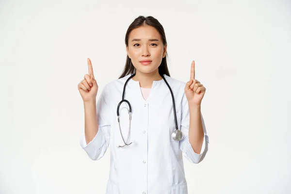 Usmívající se asijská lékařka, ukazuje prsty nahoru a dívá se do kamery, poskytují zdravotní informace, bílé pozadí — Stock fotografie