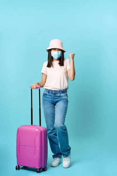 Nadšené děvče, asijský turista v lékařské masce, zavazadlo, celovečerní záběr, cestování do zahraničí na dovolené během covid-19, modré pozadí — Stock fotografie