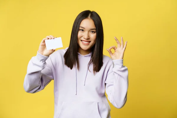 Imagem da menina coreana mostra cartão de crédito e sinal ok, conceito de compras, recomendando loja, fundo amarelo — Fotografia de Stock