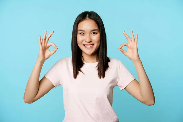 No hay problema. sonriente asiático mujer mostrando ok signos, aprobación gesto, complacido y satisfecho, de pie sobre azul fondo — Foto de Stock
