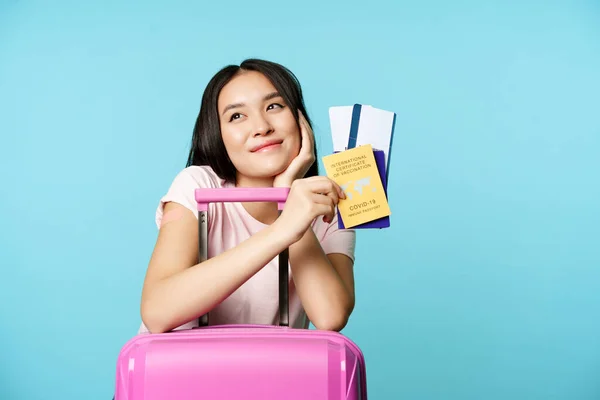 Zasněný asijský turista, usmívající se a přemýšlející, držící mezinárodní očkovací průkaz covid-19, dva lístky a pas, stojící s vhodnou péčí na výlet, modré pozadí — Stock fotografie