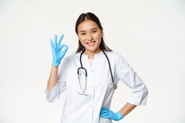 Sorrindo asiático feminino médico, enfermeira em luvas de borracha e médico uniforme mostra ok sinal, aprova e gosta smth bom, de pé sobre fundo branco — Fotografia de Stock