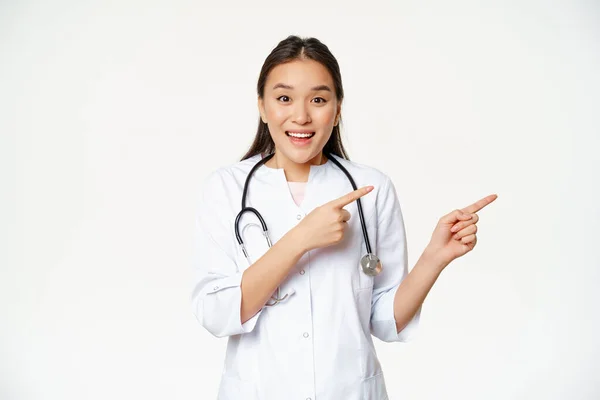 Heyecanlı gülümseyen hemşire, tıbbi üniformalı Asyalı doktor sağı gösteriyor, büyük promosyon gösteriyor, klinikte indirimler var, beyaz arka planda duruyor. — Stok fotoğraf
