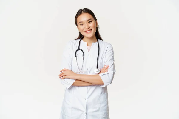 Asiatisk kvinnlig läkare, läkare i medicinsk uniform med stetoskop, kors armar på bröstet, leende och ser ut som professionell, vit bakgrund — Stockfoto