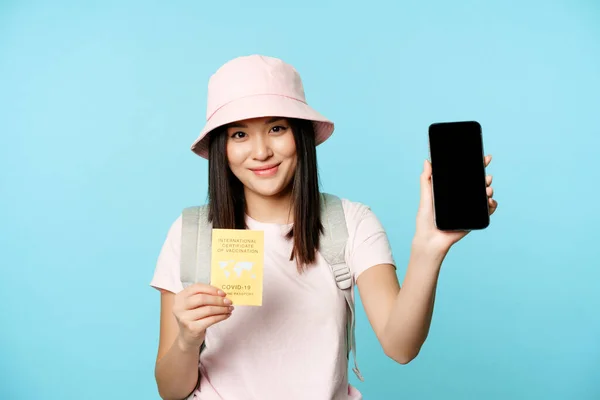 Leende asiatisk flicka visar internationella covid-19 vaccinationsintyg, hälsopass på smartphone skärm, turism app för resenärer, står över blå bakgrund — Stockfoto