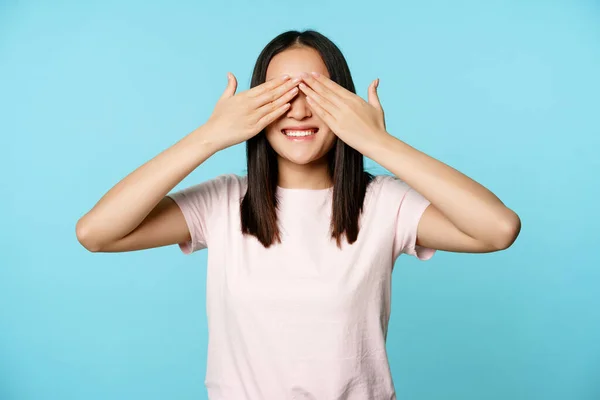 Boldog mosolygós ázsiai lány takarja fülek és várja a meglepetés, áll a póló felett kék háttér — Stock Fotó