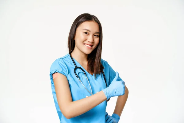 Usmívající se asijská lékařka, zdravotní sestra v drhnutí ukazuje očkovanou ruku, chřipku nebo kovid-19 vakcinační kampaň, ukazující palce nahoru, bílé pozadí — Stock fotografie