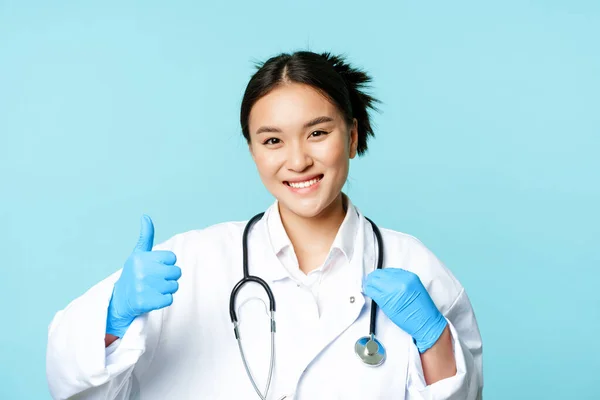 Sorrindo asiático mulher terapeuta, médico com estetoscópio mostra polegares para cima, de pé sobre fundo azul — Fotografia de Stock